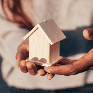 Person holding a small wooden house model for home inspection.