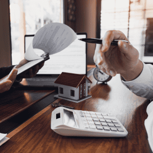 Person reviewing home documents with a calculator and model house on desk.
