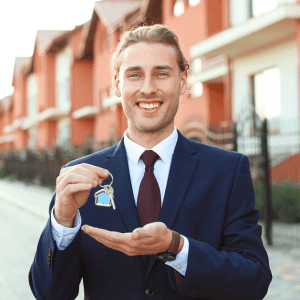 Happy man in suit holding keys in front of residential buildings.