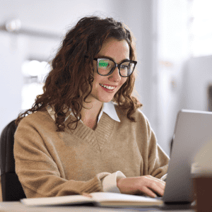 Woman with glasses smiling while working on her laptop in an office setting.