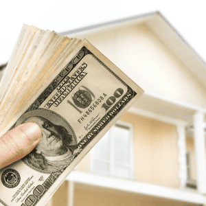 Hand holding cash in front of a house, selling property with repairs needed.